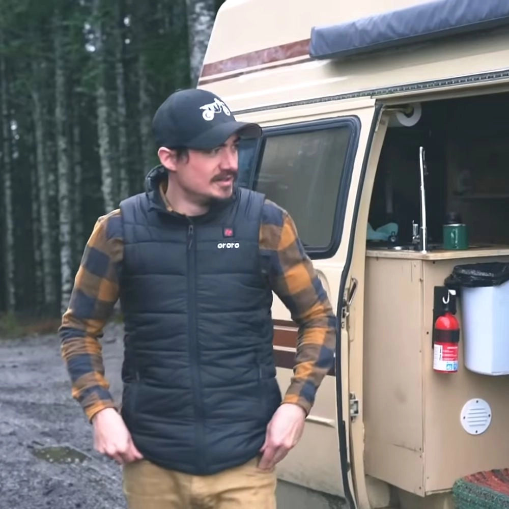 A man wearing an ororo vest standing next to a van in a forested area